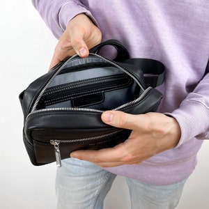 May include: A black leather zippered pouch with a silver zipper pull. The pouch is open and the inside is lined with grey fabric. There is a smaller zippered pocket inside the pouch.