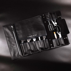 May include: Black leather tool roll with various stainless steel tools, including knives, scissors, and a whisk. The roll is open, revealing the tools in their designated pockets. The text "IN BORSHCH WE TRUST" is visible.