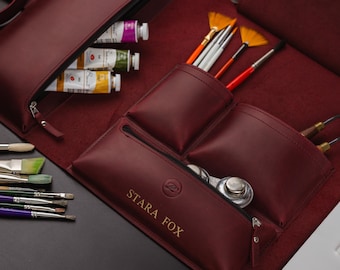 May include: A burgundy leather artist's roll-up case, open to display art supplies. The case holds paint tubes, brushes, and palette knives. The text "STARA FOX" is visible on the front of the case.