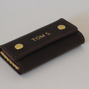 May include: A brown leather key holder with gold accents and the name "TOM S." embossed on the front.