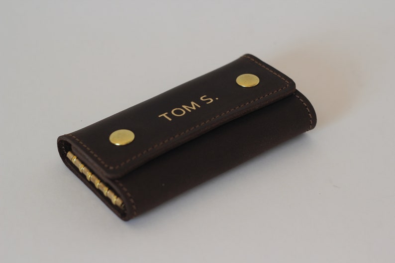 May include: A brown leather key holder with gold accents and the name "TOM S." embossed on the front.