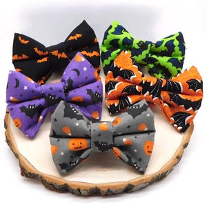 May include: Five fabric bow ties featuring Halloween-themed patterns. One is black with orange bats, one is lime green with dark bats, one is purple with bats and pumpkins, one is orange with black bats, and one is gray with pumpkins and bats.