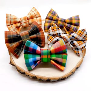May include: A collection of five fabric bow ties featuring various plaid patterns in orange, yellow, purple, brown, white, blue, and green. Each bow tie has a central fabric band and is arranged in a cluster, showcasing distinct color combinations.