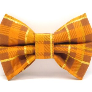 May include: A plaid bow tie in shades of orange and brown with gold accents. The bow tie has a classic shape and is made of fabric. The pattern is a grid of intersecting lines.