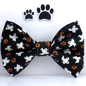 May include: A black bow tie featuring a repeating pattern of white ghosts and orange jack-o'-lanterns. Above the bow tie are two separate paw print shapes, each with a black center and a white border, matching the Halloween-themed fabric design.