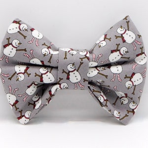 May include: A gray bow tie with a repeating pattern of cartoon snowmen. The snowmen have white bodies, brown stick arms, and wear red scarves and striped pink and white legs. The bow tie is a festive accessory.