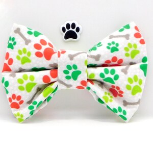 Puede incluir: Una pajarita blanca para perros con un estampado de huellas y huesos naranjas, verdes y grises. Un pequeño dije de huella está al lado de la pajarita. El accesorio es para mascotas.
