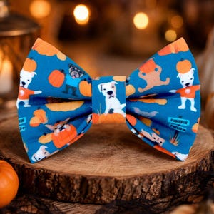 May include: A blue bow tie featuring a repeating pattern of white dogs, orange pumpkins, and a small sign that reads Pumpkin Patch. The fabric has a folded, structured shape with a central band holding the bow together in a classic style.