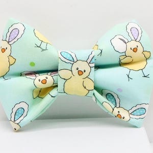 May include: A light blue bow tie with a repeating pattern of yellow chicks and white bunnies with blue ears. The bow tie is made of fabric and is suitable for Easter or spring celebrations.