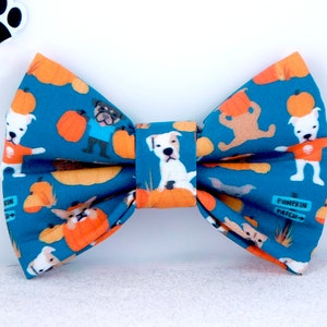 May include: A teal fabric bow tie featuring a repeating pattern of white dogs, orange pumpkins, and small blue signs that read Pumpkin Patch. The bow tie has a central fabric knot and a structured shape with folded edges throughout the design.