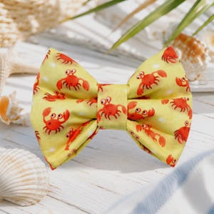 May include: A yellow bow tie featuring a repeating pattern of small, bright red cartoon crabs with large eyes and raised claws. The fabric has a soft, matte texture and the design is evenly distributed across the entire surface of the bow tie.