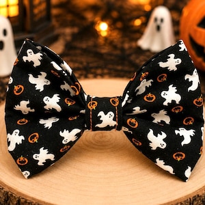 May include: A black bow tie featuring a repeating pattern of white ghosts and small orange jack-o'-lanterns. The fabric has a central knot detail with visible orange stitching, and the bow tie is shaped with two symmetrical wings.