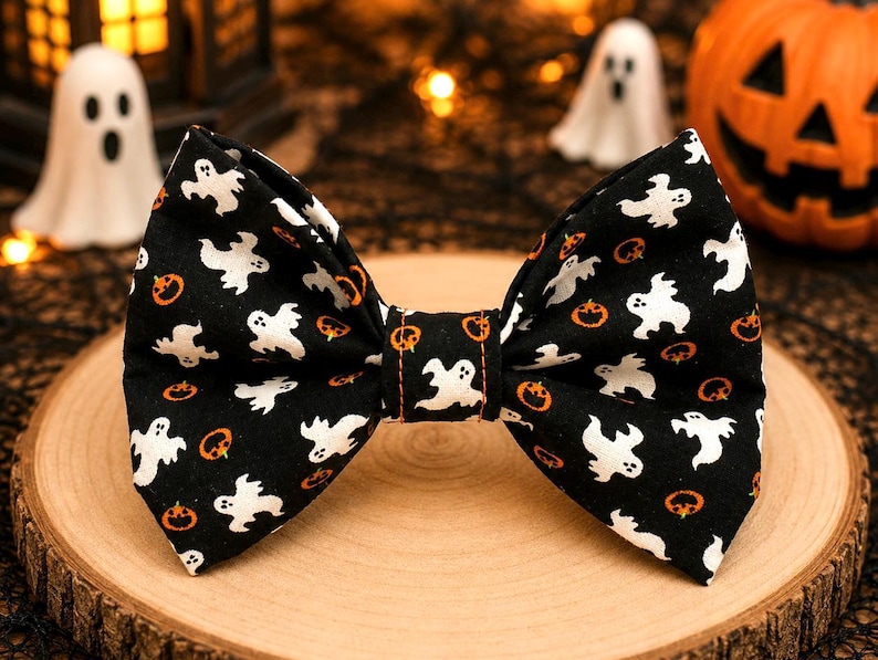 May include: A black bow tie featuring a repeating pattern of white ghosts and small orange jack-o'-lanterns. The fabric has a central knot detail with visible orange stitching, and the bow tie is shaped with two symmetrical wings.