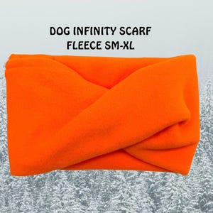 May include: Bright orange fleece dog infinity scarf, size SM-XL. The scarf is folded and twisted, creating a layered appearance. The text "DOG INFINITY SCARF FLEECE SM-XL" is printed above the scarf. The background features a snowy forest.