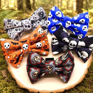May include: Five bow ties featuring various patterns including skulls, spiders, bats, candy corn, and grim reapers. The bow ties are colored in grey, blue, orange, black, and white, each with a central fabric knot and a black attachment tab.
