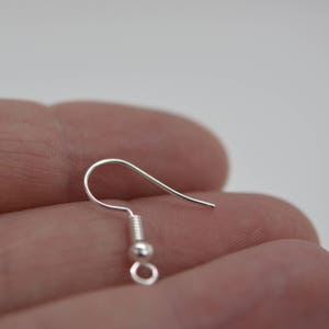 May include: A close-up of a silver-colored earring hook. The hook is curved with a small loop at the bottom and a coiled section above a small bead. The earring hook is held in a hand against a white background.