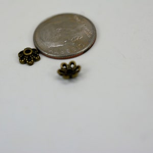 May include: Two small, gold-colored metal flower-shaped bead caps, next to a US dime for size comparison.