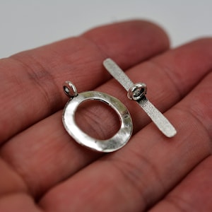 May include: A silver-colored toggle clasp, featuring a textured bar and an oval ring. The clasp is designed for jewelry making and has a rustic, antique appearance. The ring measures approximately 1 inch in diameter.