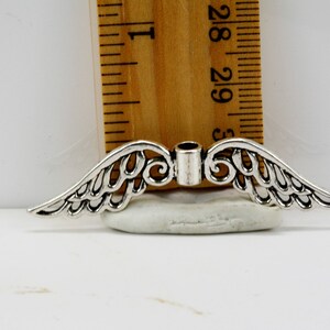May include: Silver-toned metal angel wings charm with a hole in the center for attaching to jewelry or other crafts. The wings are detailed with intricate designs and measure approximately 1.5 inches wide.