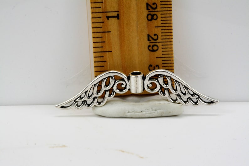 May include: Silver-toned metal angel wings charm with a hole in the center for attaching to jewelry or other crafts. The wings are detailed with intricate designs and measure approximately 1.5 inches wide.