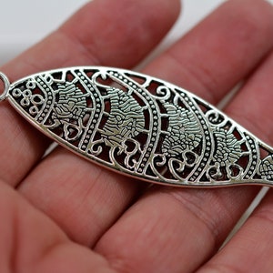 May include: Silver-colored, openwork fish pendant with intricate floral and scroll designs. The pendant has a small loop at the top for attaching to a chain or cord. The fish's tail is detailed with a fin shape. A decorative element for jewelry making.