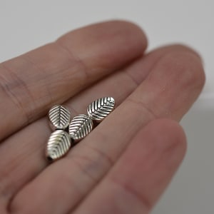 May include: Close-up of several small, silver-colored leaf-shaped beads. Each bead has detailed veining and a slightly rounded shape. The beads are held in an open hand against a plain white background.