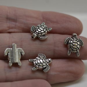 May include: Four small, silver-coloured turtle charms. Each charm has a detailed shell pattern and is designed with legs, a head, and a tail. The charms are displayed against a neutral background.