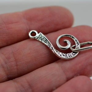 May include: Silver-toned metal charm shaped like a treble clef with an ornate design. The charm has a loop at the top for attaching to a necklace or bracelet.