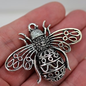 May include: A detailed silver-colored bee pendant. The bee features intricate openwork wings and body patterns. The pendant is held in a hand, showcasing its size and design. Ideal for jewelry making or as a unique charm.