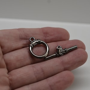 May include: A silver-toned toggle clasp, featuring a circular ring and a bar with decorative detailing. The clasp is held in a hand against a white background. This jewelry finding is used for necklaces and bracelets.