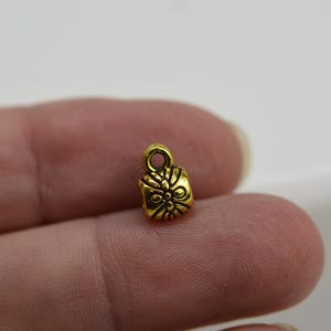 May include: A small, gold-colored charm with a floral design. The charm has a loop at the top for attaching to a necklace or bracelet. The charm is approximately 0.5 inches in height and width, and is made of metal.