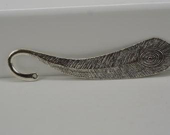May include: Silver-toned metal bookmark with a curved hook and a feather design. The feather has a textured surface and a circular pattern at the tip.