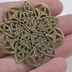 May include: A detailed, antique brass-colored metal filigree flower. The openwork design features a central flower shape with six petals, each with intricate cut-out patterns. The overall diameter is approximately 2 inches.