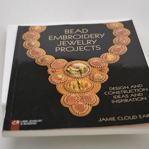May include: A softcover book titled Bead Embroidery Jewelry Projects by Jamie Cloud Eakin. The black cover features a graphic illustration of a beaded necklace with gold and brown tones, along with the text Design and Construction, Ideas and Inspiration.