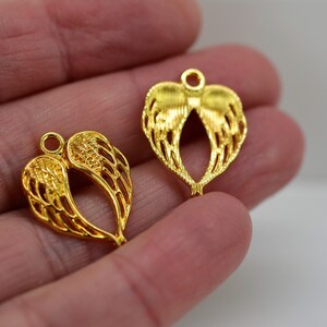 K&ouml;nnte beinhalten: Zwei goldfarbene Metall-Engel Fl&uuml;gel Charms. Die Charms sind in Form von Fl&uuml;geln mit einer herzf&ouml;rmigen &Ouml;ffnung in der Mitte. Die Charms sind auf einer Hand.