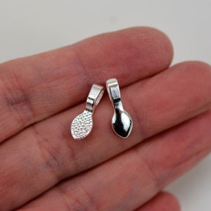 May include: Two small, silver-colored jewelry bails. One bail has a textured, dotted surface on the teardrop-shaped base, while the other has a smooth, polished surface. Both have a small loop at the top for attaching to a chain or other jewelry component.