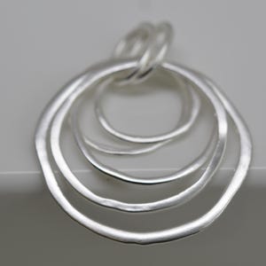 May include: A silver pendant featuring four interconnected, irregularly shaped rings. The rings are of varying sizes, with a textured, matte finish. The pendant is suspended from a small loop at the top, suggesting it's designed to be worn as jewelry.