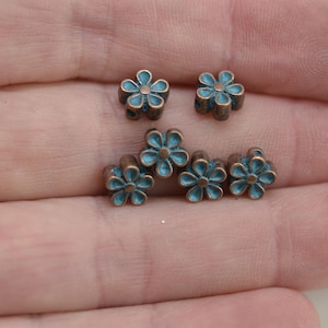 May include: A collection of six small, copper-toned metal flower charms with blue enamel centers. The charms are arranged on a hand.