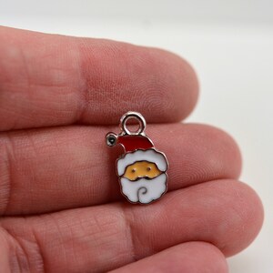May include: Silver charm with a white and red Santa Claus design. The Santa Claus has a white beard, a red hat, and a yellow face.