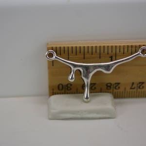 Puede incluir: Encanto de metal plateado en forma de l&iacute;quido que gotea con dos anillos de salto. El encanto mide aproximadamente 2,5 cm de largo y se muestra en una regla.