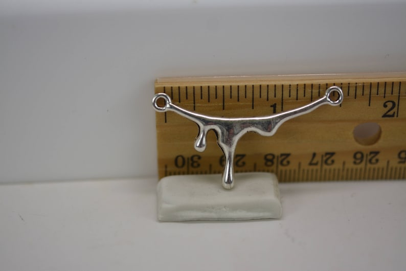 Puede incluir: Encanto de metal plateado en forma de l&iacute;quido que gotea con dos anillos de salto. El encanto mide aproximadamente 2,5 cm de largo y se muestra en una regla.