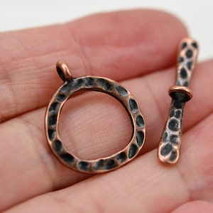May include: A hammered copper toggle clasp with a round ring and a straight bar. The clasp has a dark patina finish.