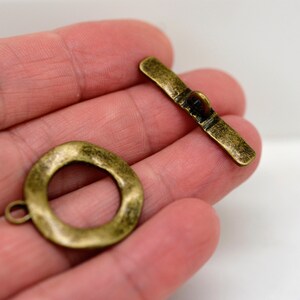 May include: Antique brass toggle clasp set. The set includes a textured, round ring and a bar with a central toggle. The clasp has a rustic, aged appearance, suggesting vintage or handmade jewelry components.