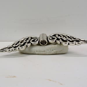 May include: Silver angel wing charm with a clear crystal in the center. The charm is designed to be used as a pendant or a bead for jewelry making.