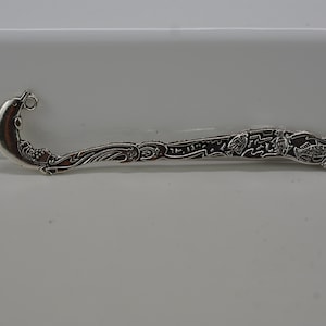May include: A silver-colored bookmark with a dolphin design at the top. The bookmark has an elongated shape with detailed carvings of marine life and floral patterns. The dolphin has a small ring attached to its fin.