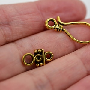 Peut inclure: Deux fermoirs en m&eacute;tal dor&eacute; pour la fabrication de bijoux. Un fermoir est une simple boucle avec un crochet, l'autre est un design d&eacute;coratif et orn&eacute; avec deux boucles.