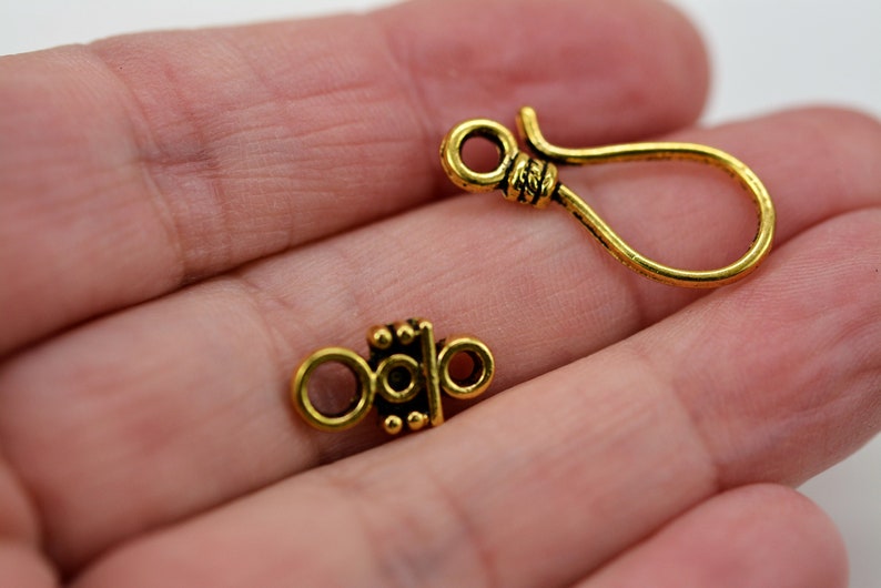Peut inclure: Deux fermoirs en m&eacute;tal dor&eacute; pour la fabrication de bijoux. Un fermoir est une simple boucle avec un crochet, l'autre est un design d&eacute;coratif et orn&eacute; avec deux boucles.
