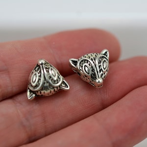 May include: Two silver fox head beads with intricate details. The beads are triangular in shape and have a textured surface.