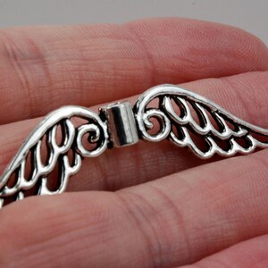 May include: Silver-toned metal angel wings charm with a delicate, intricate design. The wings are connected by a small metal loop.