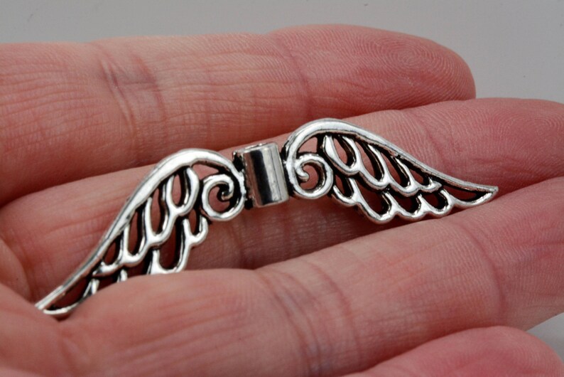 May include: Silver-toned metal angel wings charm with a delicate, intricate design. The wings are connected by a small metal loop.